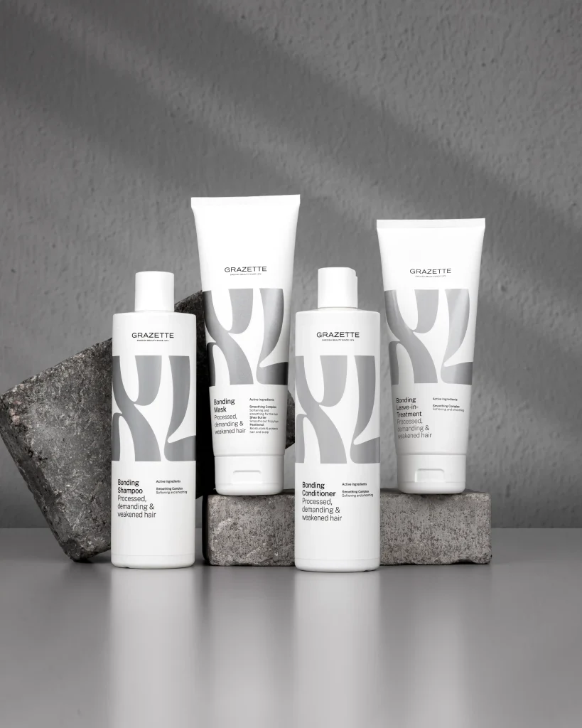 En gruppbild på hårvårdsserien Grazette XL Bonding (shampoo, mask, conditioner och leave-in) uppställda på gråa stenblock. Bilden går i en monokrom gråskala med mjukt infallande ljus mot en strukturerad betongvägg.